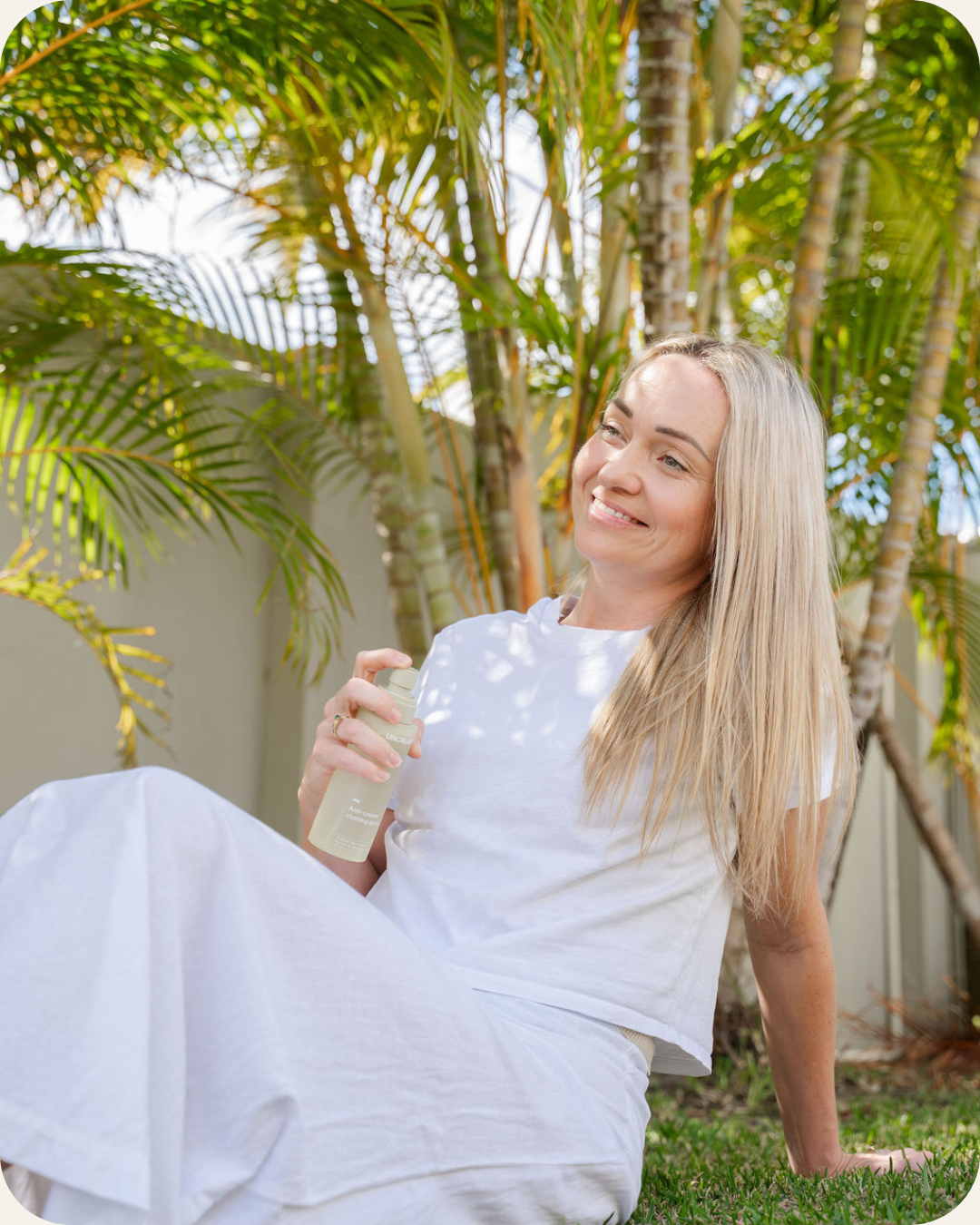 A woman with long blonde hair sits on grass outdoors among palm trees, wearing a white t-shirt and white pants while holding an Uncrumpled spray bottle.