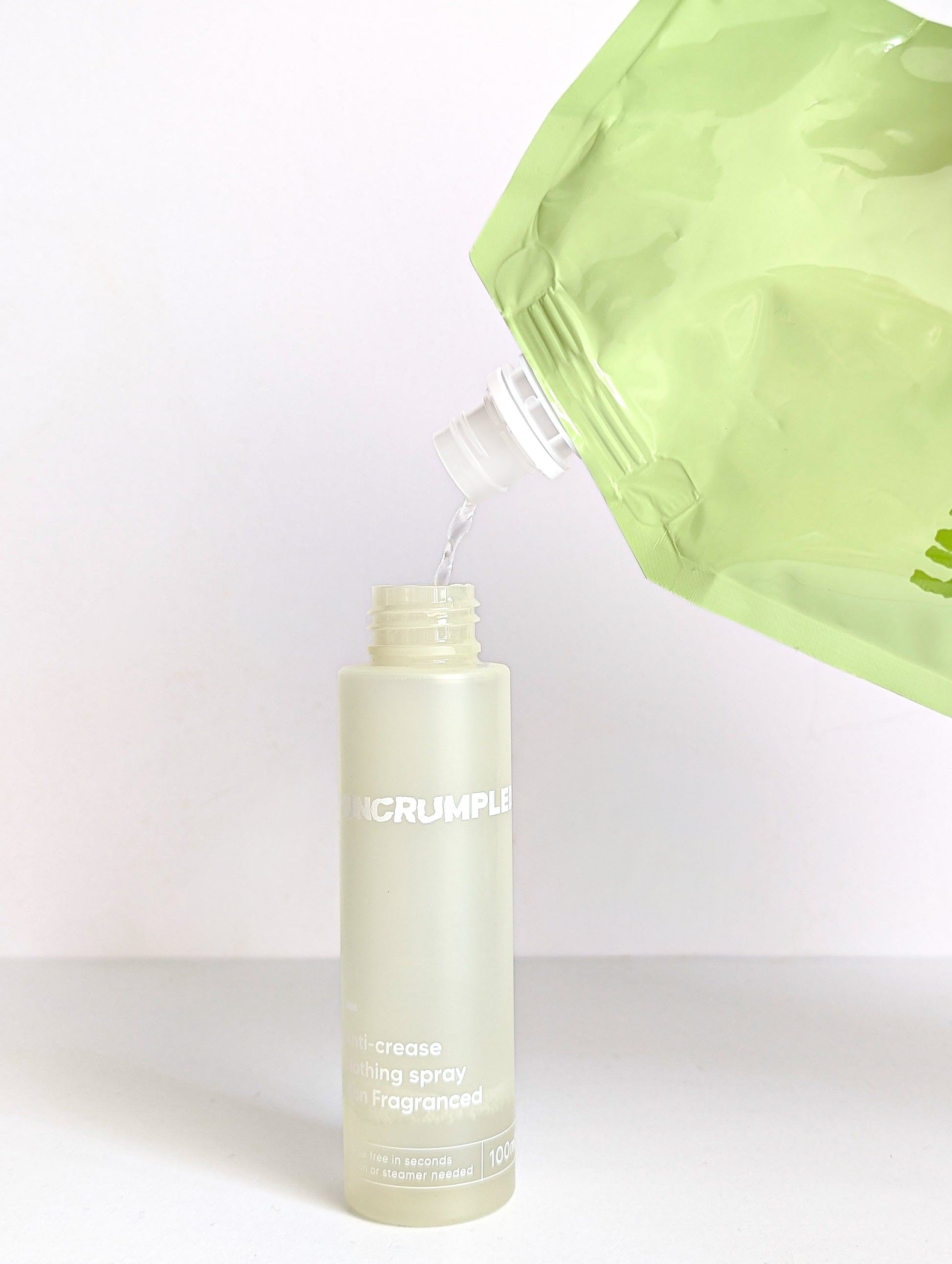 Travel-sized Uncrumpled anti-crease spray bottle with its cap and nozzle off, liquid content being poured into it from Uncrumpled refill pouch.